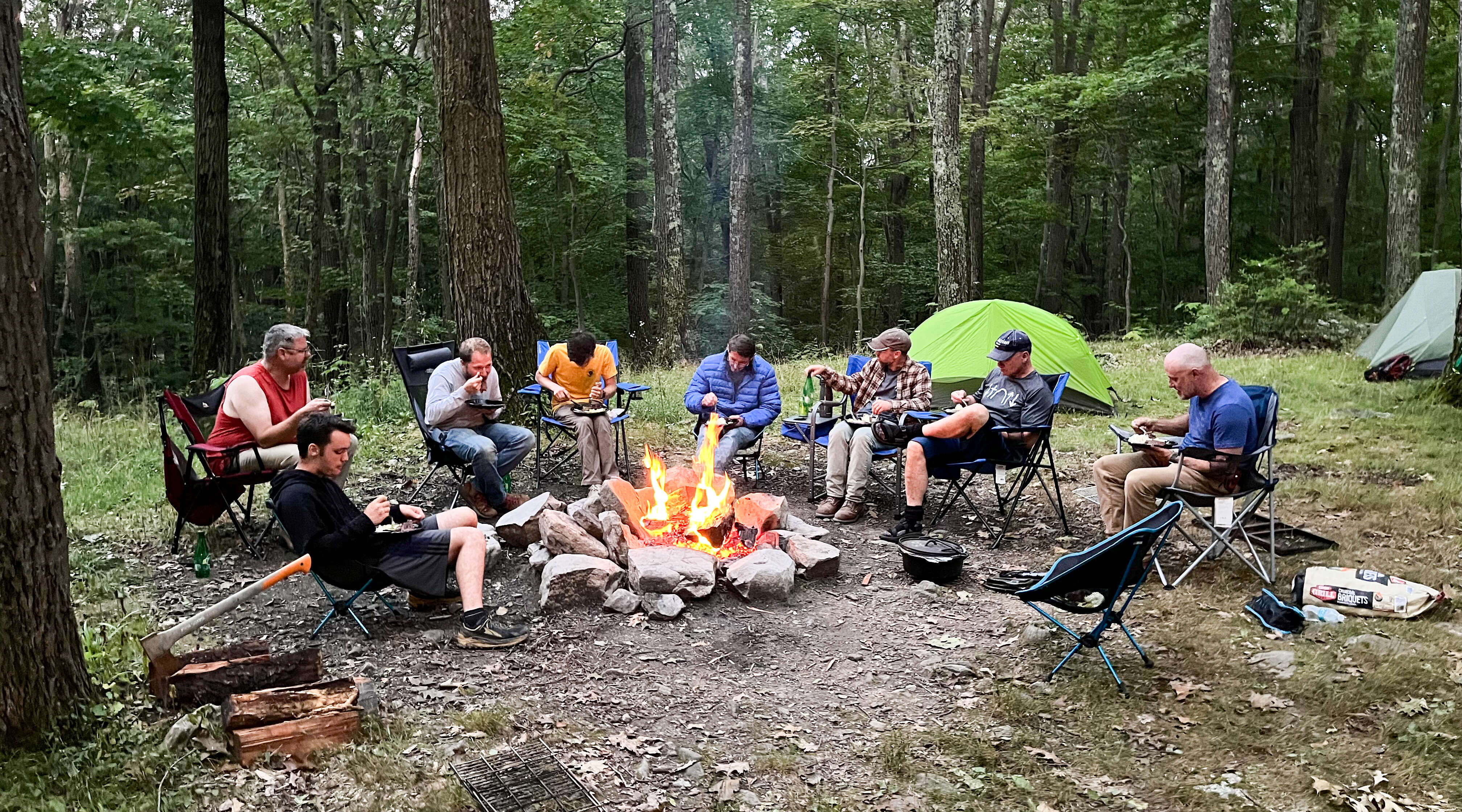 A group of guys eating around the campfire.