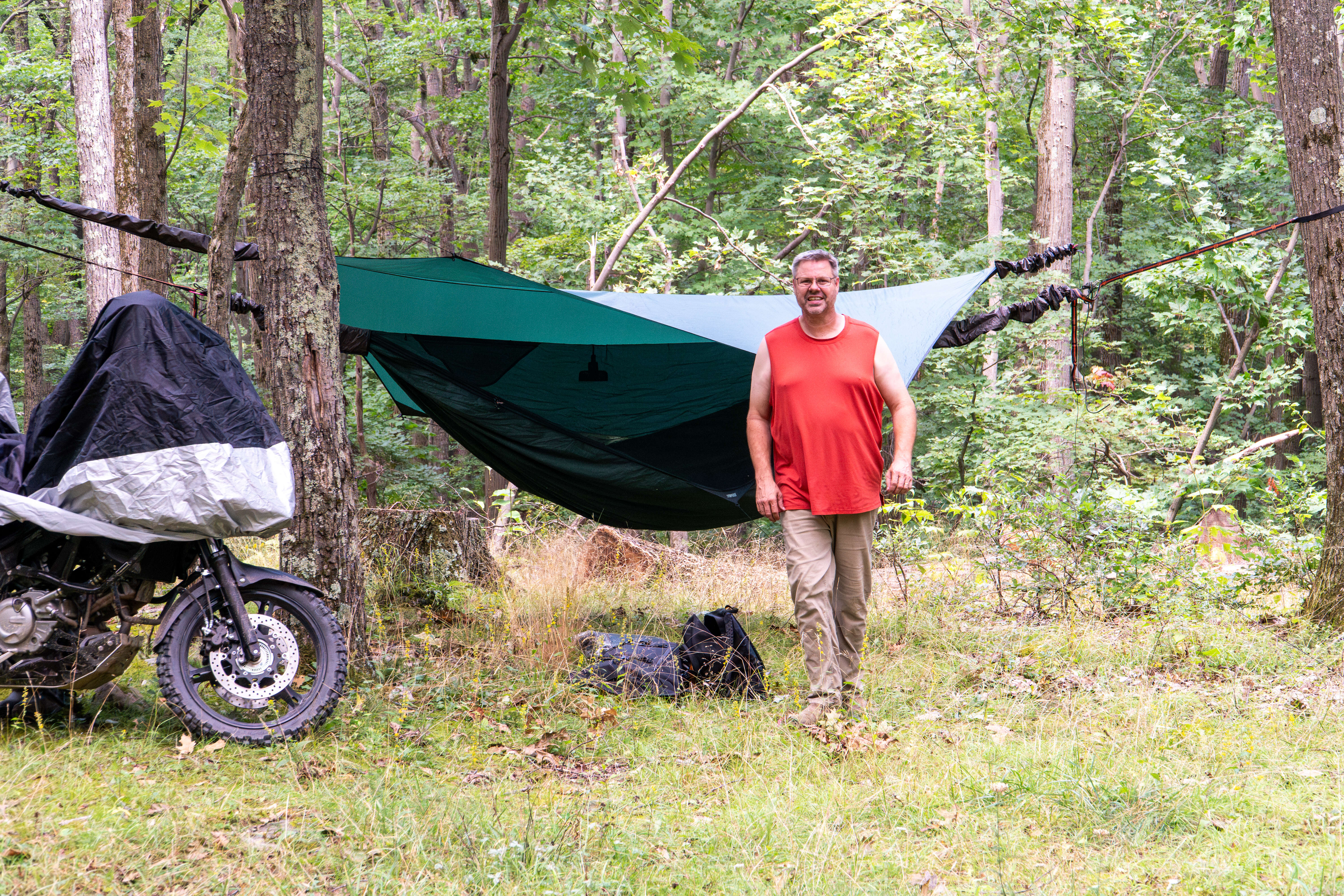 A hammock setup in the woods with a guy standing in front of it with a motorcycle off to the left.